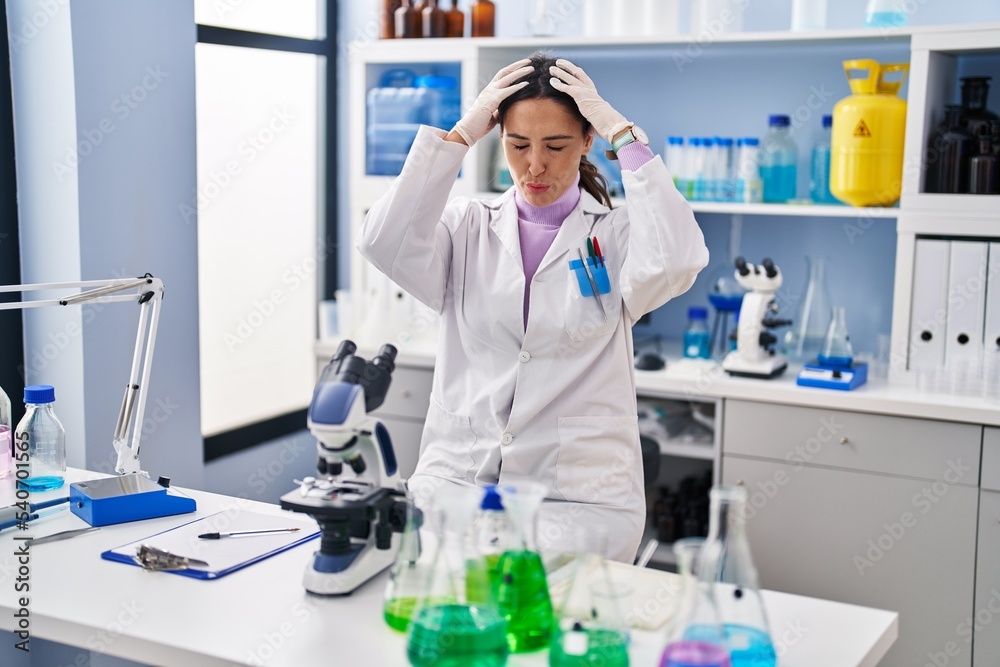 Young brunette woman working at scientist laboratory suffering from headache desperate and stressed because pain and migraine. hands on head.