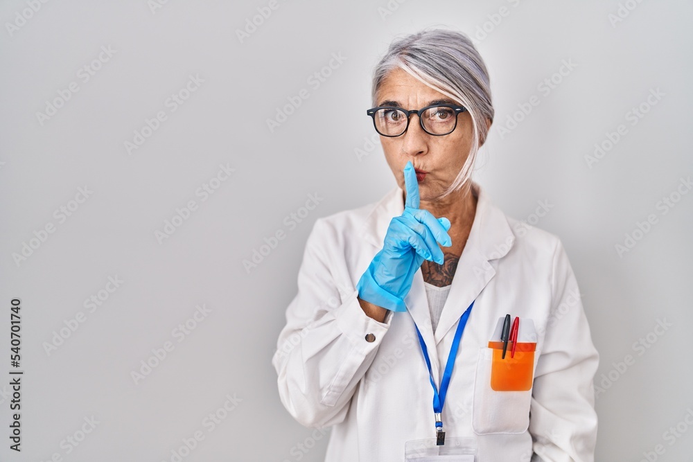 Middle age woman with grey hair wearing scientist robe asking to be quiet with finger on lips. silence and secret concept. Middle age woman with grey hair wearing scientist robe asking to be quiet with finger on lips. silence and secret concept.