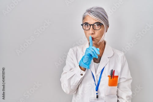 Middle age woman with grey hair wearing scientist robe asking to be quiet with finger on lips. silence and secret concept.