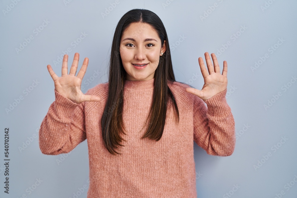Young latin woman standing over blue background showing and pointing up with fingers number ten while smiling confident and happy. Young latin woman standing over blue background showing and pointing up with fingers number ten while smiling confident and happy.