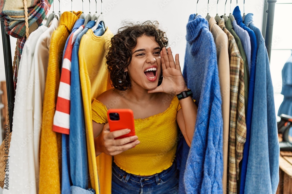 Young hispanic woman searching clothes on clothing rack using smartphone shouting and screaming loud to side with hand on mouth. communication concept.