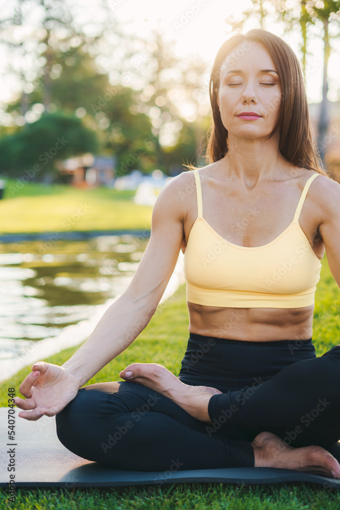Brunette woman in sportswear sitting in lotus position with eyes closed enjoying meditation on sunny morning in nature. Training workout. Healthy activity