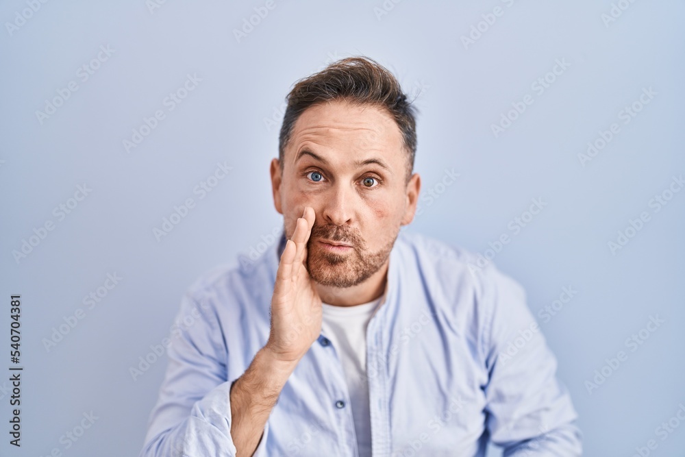 Middle age caucasian man standing over blue background hand on mouth telling secret rumor, whispering malicious talk conversation