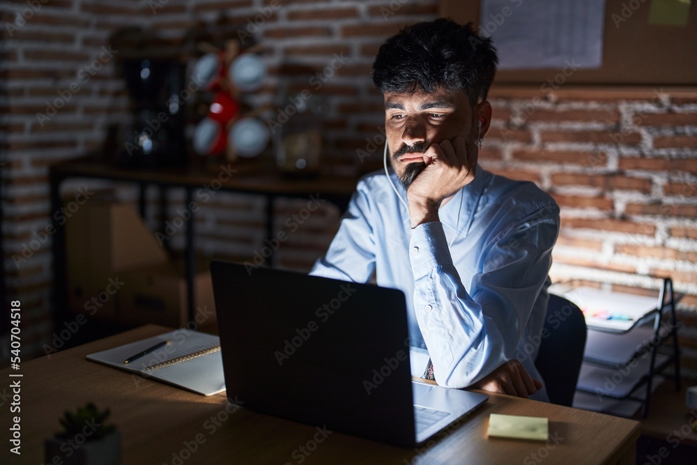 Young hispanic man with beard working at the office at night thinking looking tired and bored with depression problems with crossed arms.