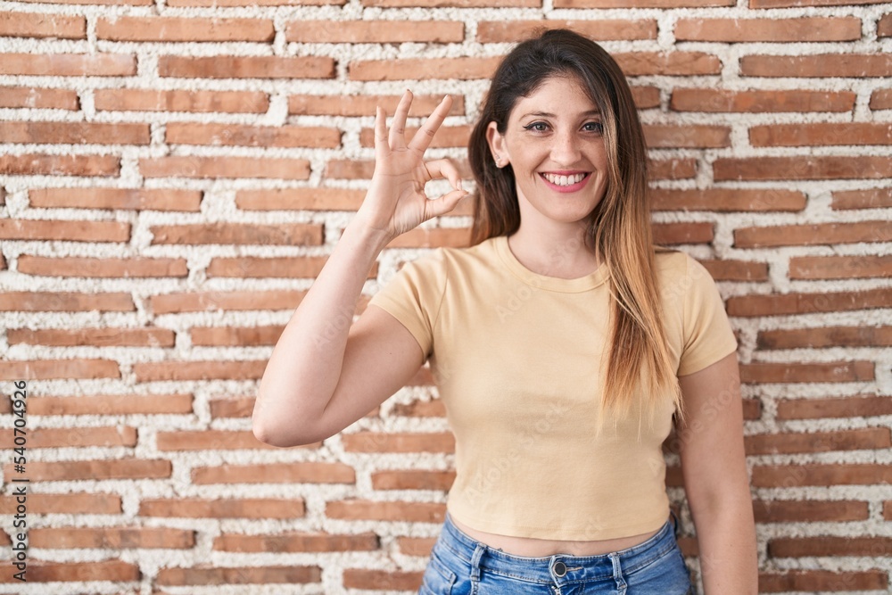 Young brunette woman standing over bricks wall smiling positive doing ok sign with hand and fingers. successful expression. Young brunette woman standing over bricks wall smiling positive doing ok sign with hand and fingers. successful expression.