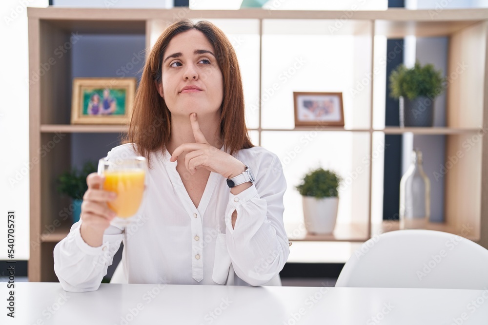 Brunette woman drinking glass of orange juice thinking concentrated about doubt with finger on chin and looking up wondering Brunette woman drinking glass of orange juice thinking concentrated about doubt with finger on chin and looking up wondering