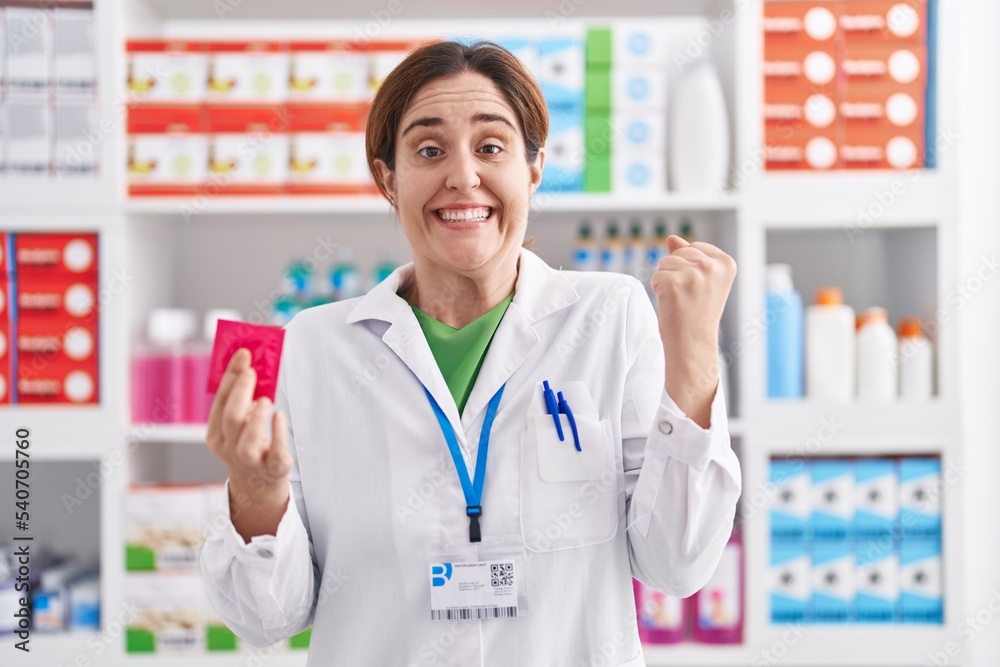 Brunette woman working at pharmacy drugstore holding condom screaming proud, celebrating victory and success very excited with raised arms