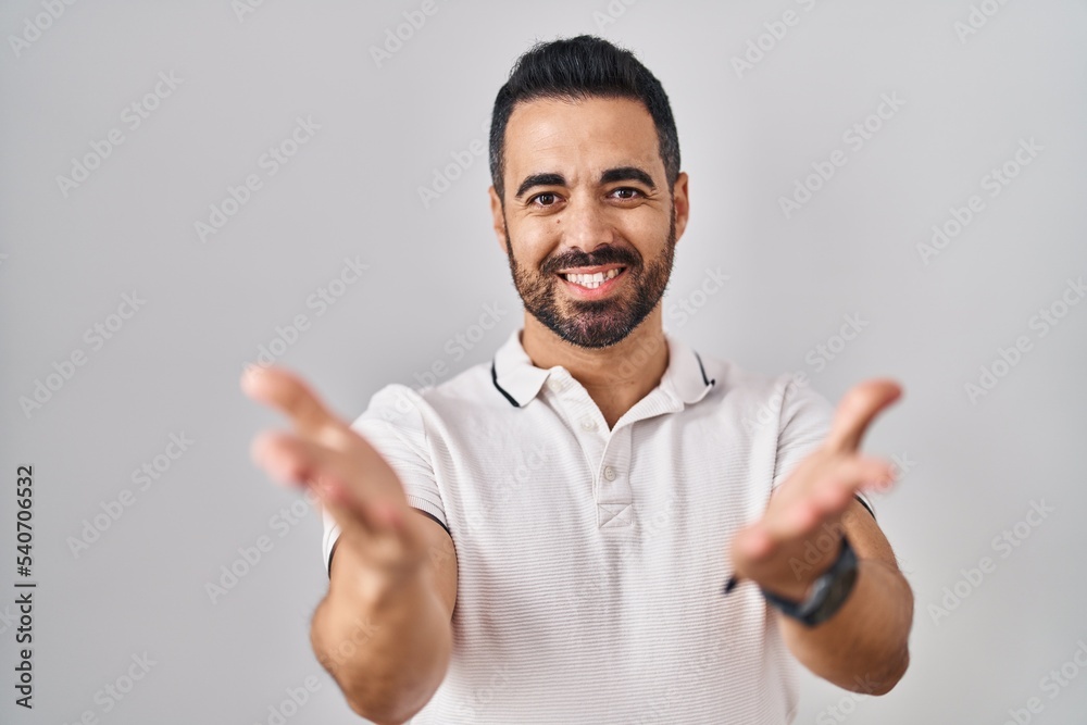 Fototapeta premium Young hispanic man with beard wearing casual clothes over white background smiling cheerful offering hands giving assistance and acceptance.