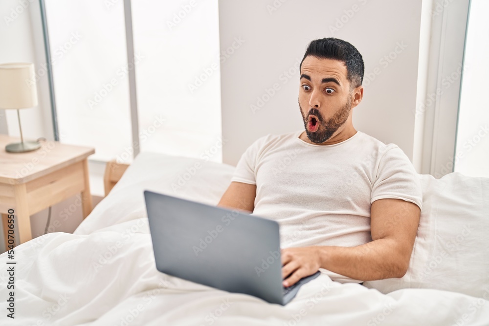 Young hispanic man with beard using computer laptop on the bed scared and amazed with open mouth for surprise, disbelief face Young hispanic man with beard using computer laptop on the bed scared and amazed with open mouth for surprise, disbelief face
