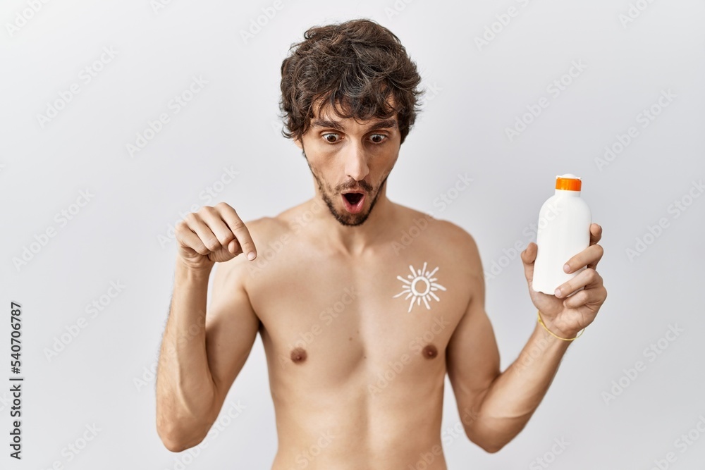 Fototapeta premium Young hispanic man standing shirtless holding sunscreen lotion pointing down with fingers showing advertisement, surprised face and open mouth