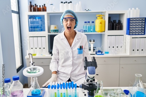 Brunette woman working at scientist laboratory angry and mad screaming frustrated and furious  shouting with anger. rage and aggressive concept.
