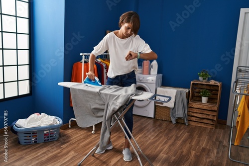 Young beautiful hispanic woman ironing clothes looking watch at laundry room
