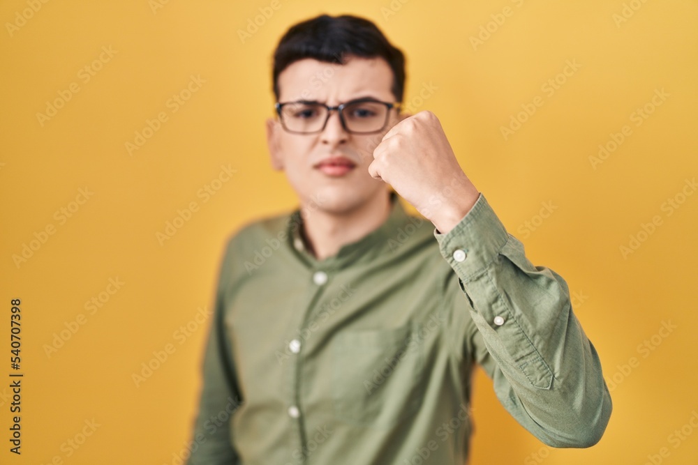 Non binary person standing over yellow background angry and mad raising fist frustrated and furious while shouting with anger. rage and aggressive concept. Non binary person standing over yellow background angry and mad raising fist frustrated and furious while shouting with anger. rage and aggressive concept.