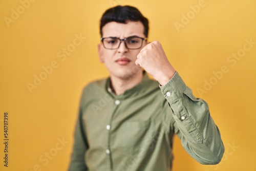 Non binary person standing over yellow background angry and mad raising fist frustrated and furious while shouting with anger. rage and aggressive concept.