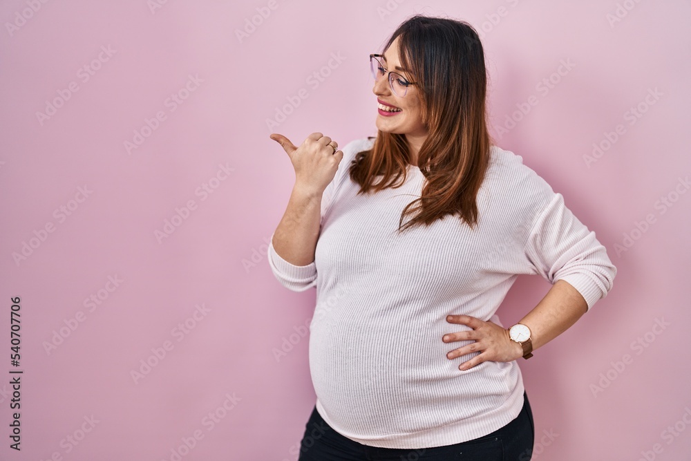 Pregnant woman standing over pink background smiling with happy face looking and pointing to the side with thumb up. Pregnant woman standing over pink background smiling with happy face looking and pointing to the side with thumb up.