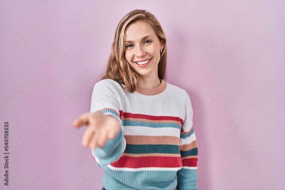 Young blonde woman standing over pink background smiling cheerful offering palm hand giving assistance and acceptance. Young blonde woman standing over pink background smiling cheerful offering palm hand giving assistance and acceptance.