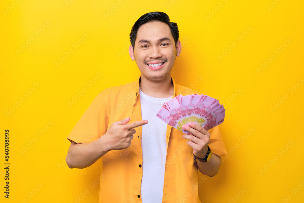 Smiling young handsome Asian man pointing fingers at money banknotes isolated on yellow background Smiling young handsome Asian man pointing fingers at money banknotes isolated on yellow background