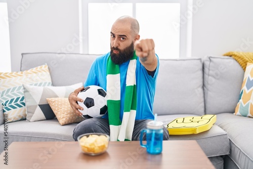 Young hispanic man with beard and tattoos football hooligan holding ball supporting team pointing with finger to the camera and to you, confident gesture looking serious © Krakenimages.com