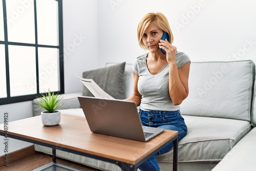 Middle age blonde woman talking on the smartphone using laptop at home