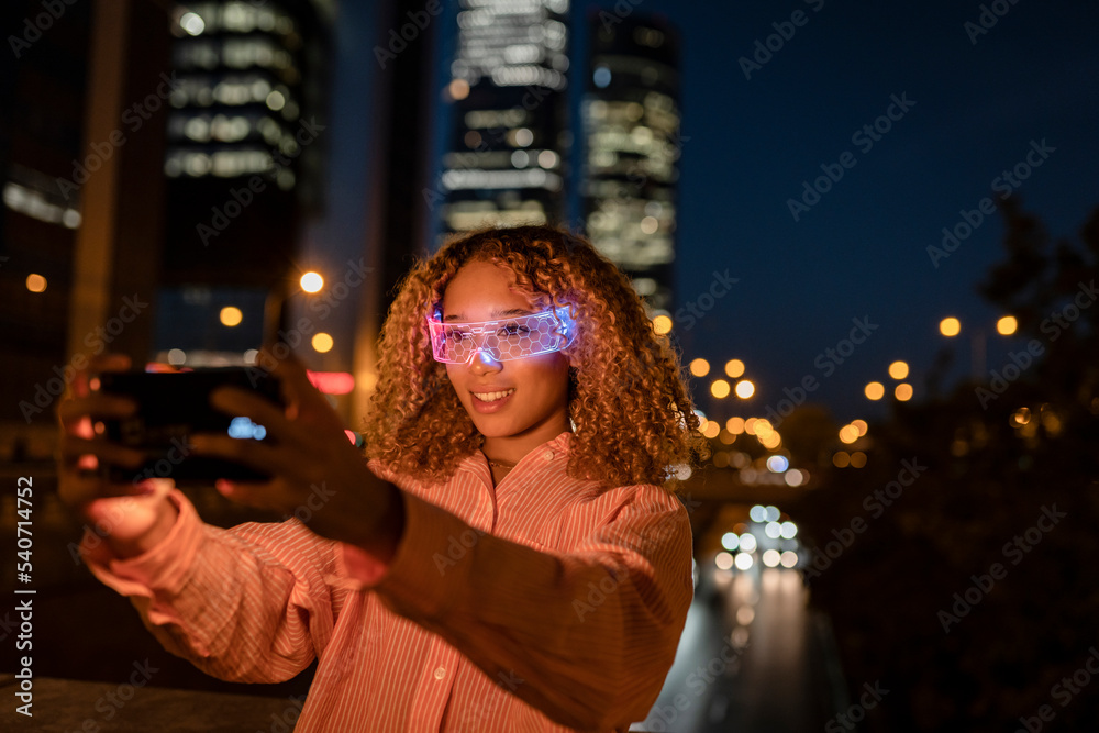 Smiling woman with glowing smart glasses taking selfie on smart phone at night Smiling woman with glowing smart glasses taking selfie on smart phone at night