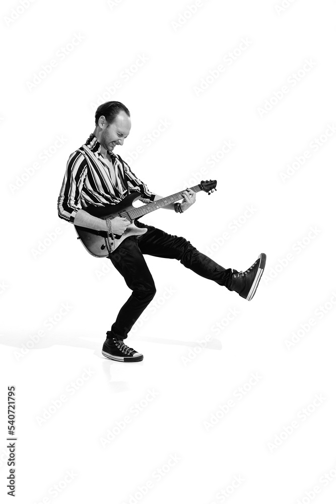 Portrait of young emotive man in stylish clothes playing guitar, posing isolated over white background. Black and white photo