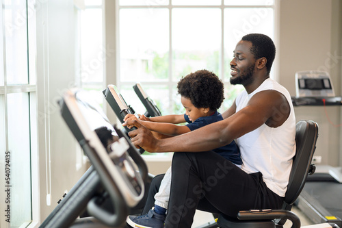African American man with child exercising on the exercise bike in indoor fitness club. Healthy and Fitness concept.