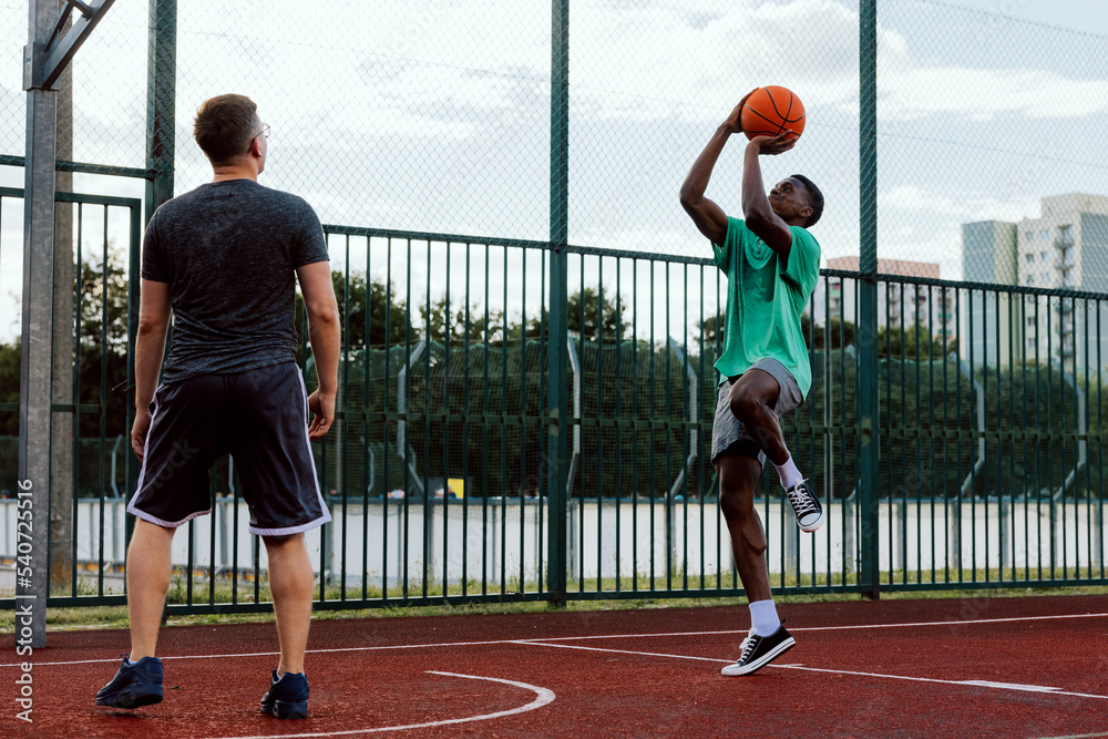 Professional basketball player with friends at basketball court showing skills training friends guys making slam dunk. Happy guys playing game outside wearing sportswear.