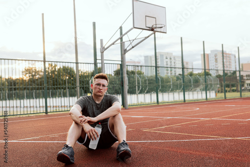 Young tired exhausted guy man in glasses wearing sportswear sitting at basketball court outside resting relaxing after training time doing exercises with company guys friends. Basketball game process.