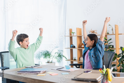 excited kids showing win gesture near notebooks and paper notes on table at home.