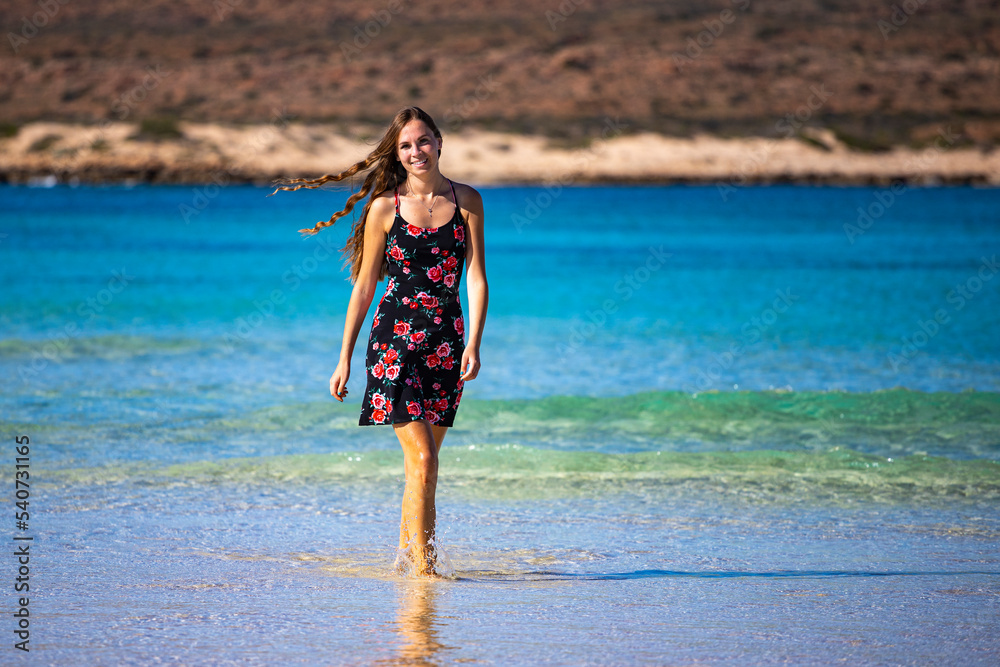beautiful long-haired girl in a dress soaks her legs in turquoise water on a paradise beach at turquoise bay in cape range national park near exmouth, western australia; beach with red cliffs in the b