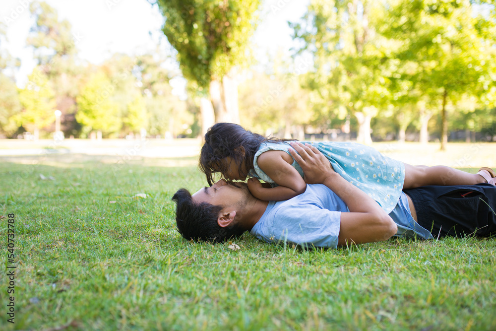 Side view of Asian dad resting with little girl on field. Happy young father lying on green grass in public park holding his daughter and kissing her. Happy childhood and summer rest together concept Side view of Asian dad resting with little girl on field. Happy young father lying on green grass in public park holding his daughter and kissing her. Happy childhood and summer rest together concept