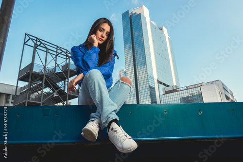 Stylish beautiful casual caucasian hip hop dancer posing sitting in blue jacket, jeans, black top and white tshirt in urban town background. Outdoor city shooting of a model photo