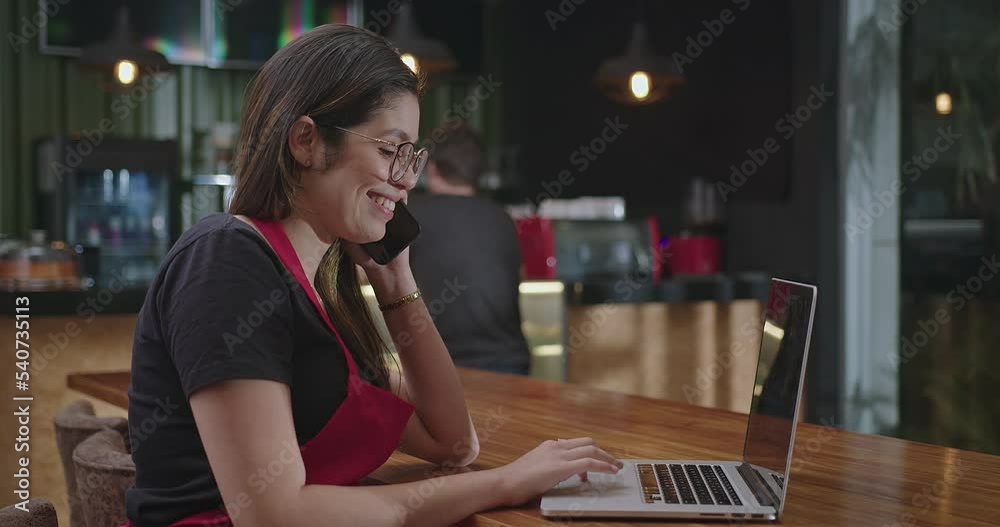 Female hispanic barista employee working in front of laptop computer taking online delivery. Young woman using technology speaking with client and multi tasking
