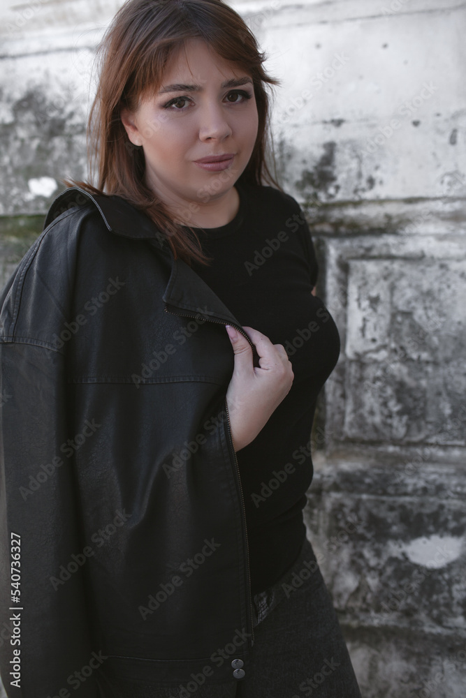 Plus size woman dressed jacket, jeans and t-shirt posing against wall in the street
