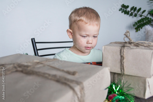 A small child with Christmas gifts is sitting at a New Year's wooden table with a tablet in his hands. Celebrating Christmas.