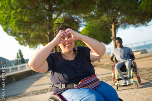 Portrait of cheerful Caucasian woman in park. Woman in wheelchair in casual clothes on summer day, showing heart sign. Boyfriend in background. Portrait, beauty, happiness concept