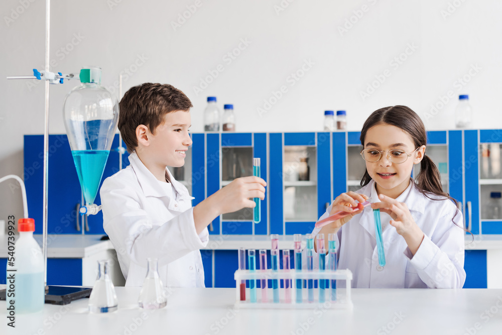 happy preteen kids in white coats looking at camera near microscope and ...