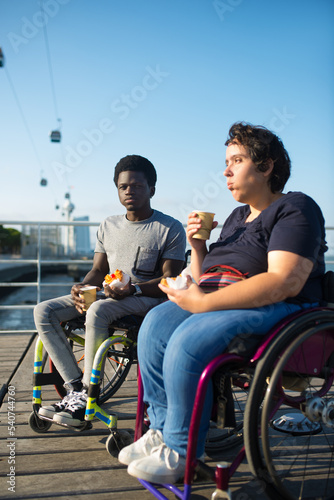 Content biracial couple drinking coffee on sunny day. African American man and Caucasian woman in wheelchairs on embankment, drinking hot beverage from cups. Snack, relationship, happiness concept