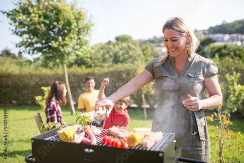 Smiling woman cooking BBQ meat and vegetables. Mid adult woman in khaki dress turning meat on grid. Family in background. BBQ, family leisure, food concept