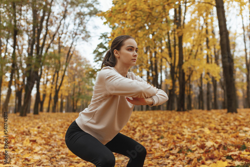 Focused fit sportswoman doing squat exercise while having fitness training in autumn park and leading healthy lifestyle 