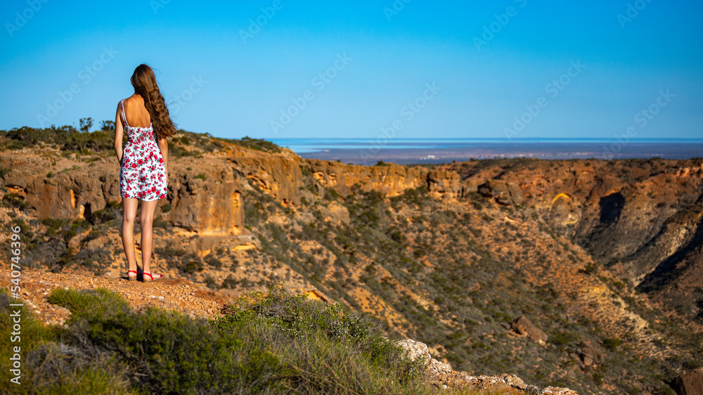 Naklejka premium a beautiful girl in a short white dress stands on top of a mountain above a massive gorge in western australia at sunset, hiking in cape range national park near exmouth