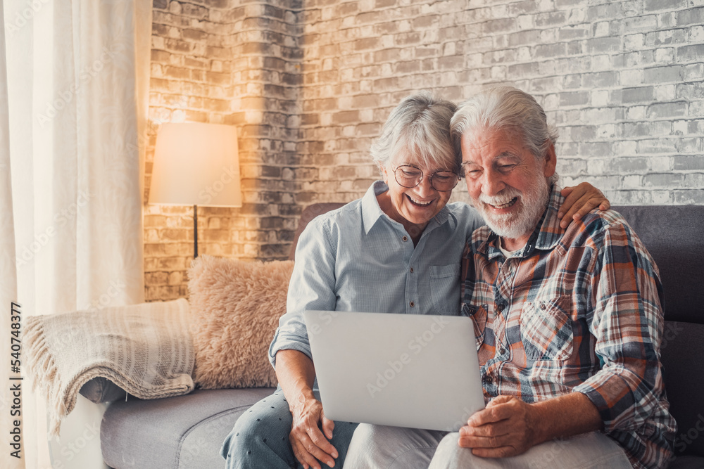 Cute couple of old people sitting on the sofa using laptop together shopping and surfing the net. Two mature people in the living room enjoying technology. Portrait of seniors laughing in love..