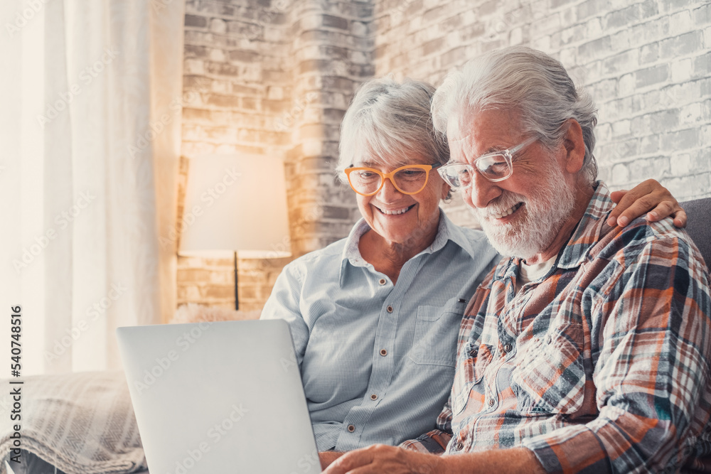 Cute couple of old people sitting on the sofa using laptop together shopping and surfing the net. Two mature people wearing eyeglasses in the living room enjoying technology. Portrait of seniors 