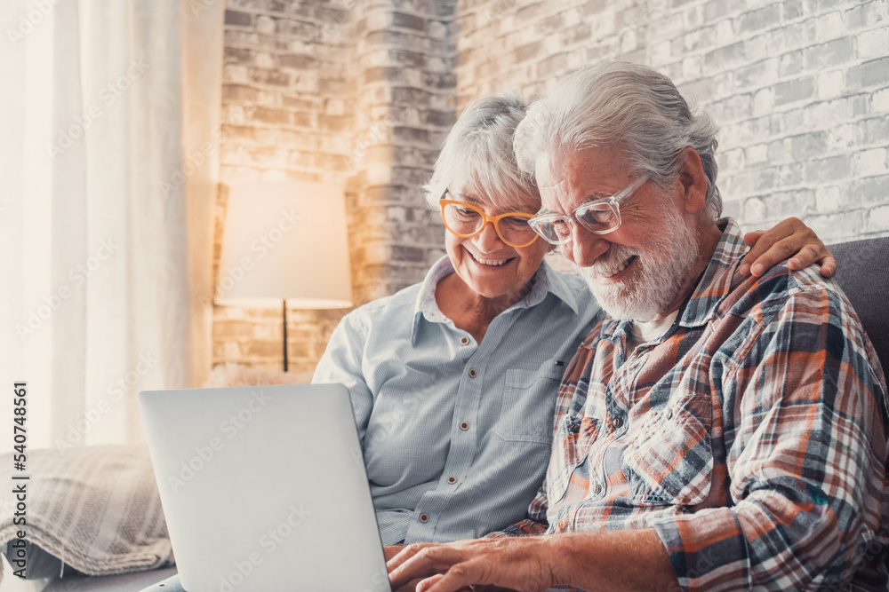 Cute couple of old people sitting on the sofa using laptop together shopping and surfing the net. Two mature people wearing eyeglasses in the living room enjoying technology. Portrait of seniors Cute couple of old people sitting on the sofa using laptop together shopping and surfing the net. Two mature people wearing eyeglasses in the living room enjoying technology. Portrait of seniors