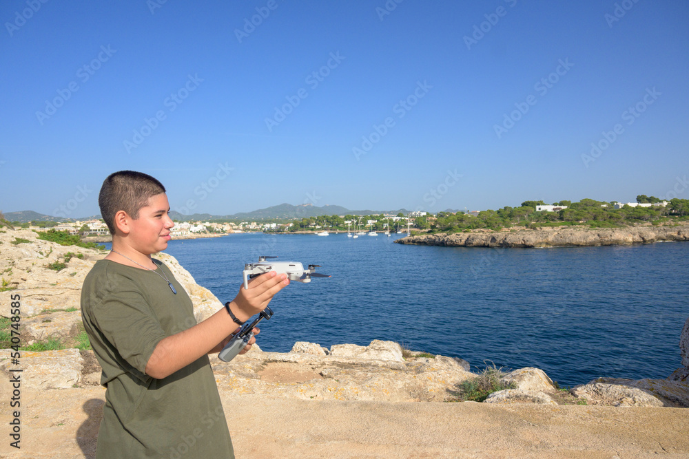 happy teenage boy, preparing and finalizing flight details for drone flight during sunny day with the mediterranean sea in the background Spain, Balearic Islands