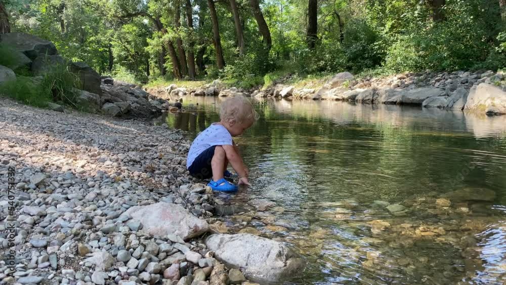 Little child playing on river bank in a summer park, 1.5 year old toddler enjoys throwing pebbles into the water. Little child playing on river bank in a summer park, 1.5 year old toddler enjoys throwing pebbles into the water.