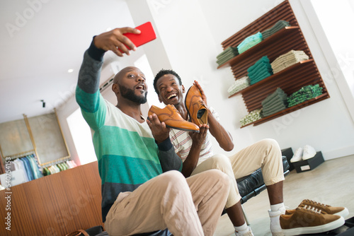 African American gay men holding modern shoes. Two smiling boyfriends sitting on bench at clothing store trying on shoes and taking selfie. LGBT couples relations, fashion choice and purchases concept photo