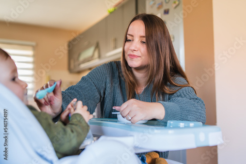 A mother is feeding her baby son in his chair during the day while baby is smiling and enjoying his meal