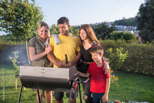 Focused father grilling meat and vegetables with family. Dark-haired man in yellow T-shirt standing near BBQ grid with little son, wife and teenage daughter. BBQ, cooking, food, family concept