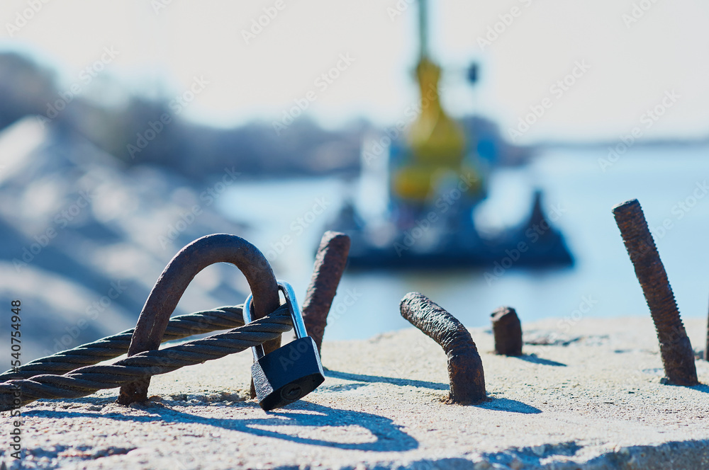 Fototapeta premium The steel cable is fixed on a reinforced concrete slab in the port. Sunny day. Simple padlock. Bracing and securing concept. Selective focus in the foreground.